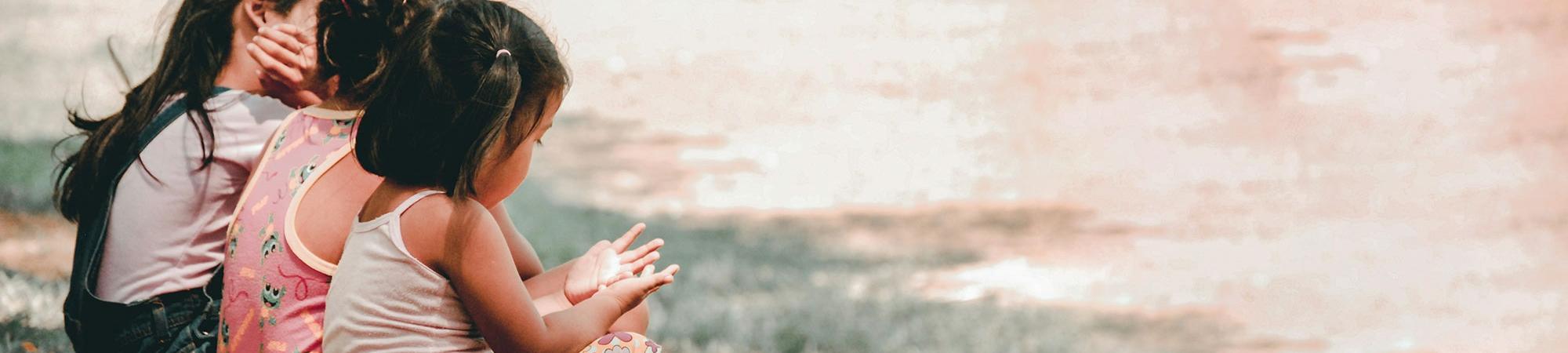 Three children sitting in the grass on the left side of the image. Photo by Charlein Gracia via UnSplash